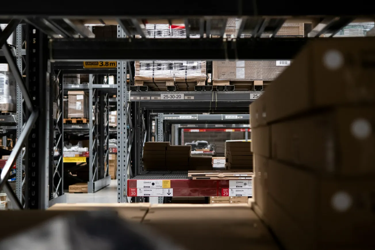 A warehouse aisle with tall metal shelves stacked with cardboard boxes and pallets. Signs with numbers hang overhead, indicating an organized and industrial atmosphere.