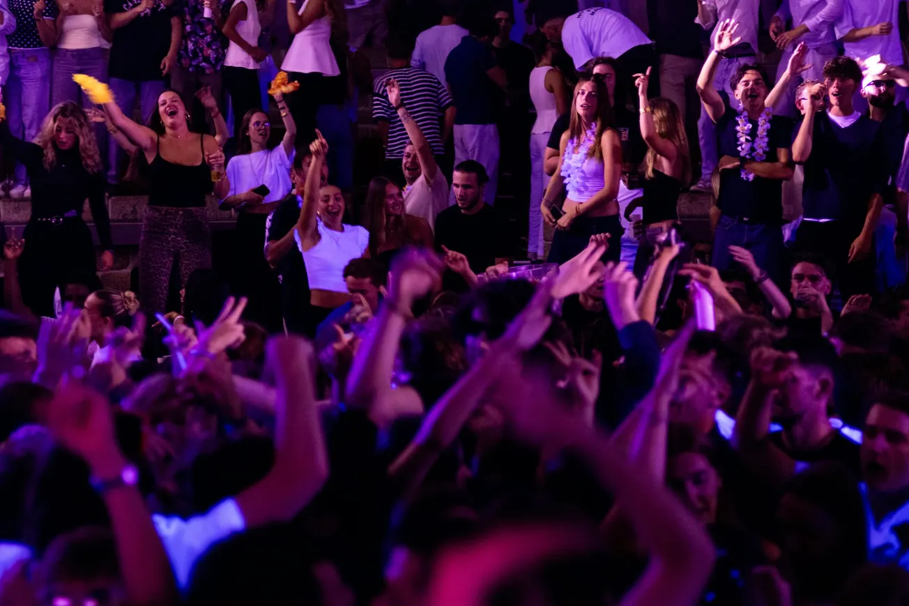 A vibrant crowd at a concert, arms raised, dancing under purple lights. People smile and cheer, conveying joy and excitement in a lively atmosphere.