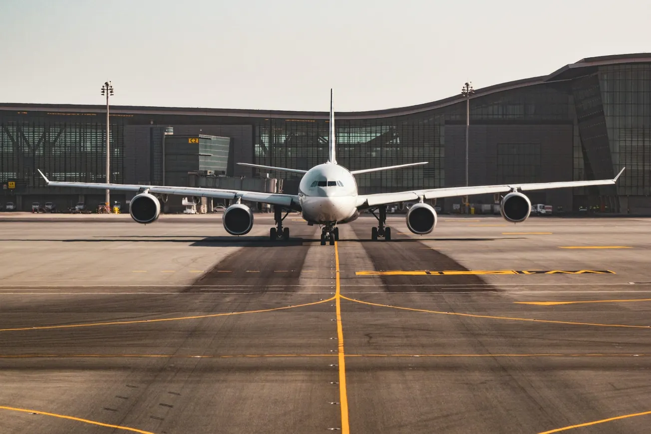 Front view of a large white airplane taxiing on a runway at an airport, with a modern glass terminal building in the background under a clear sky.