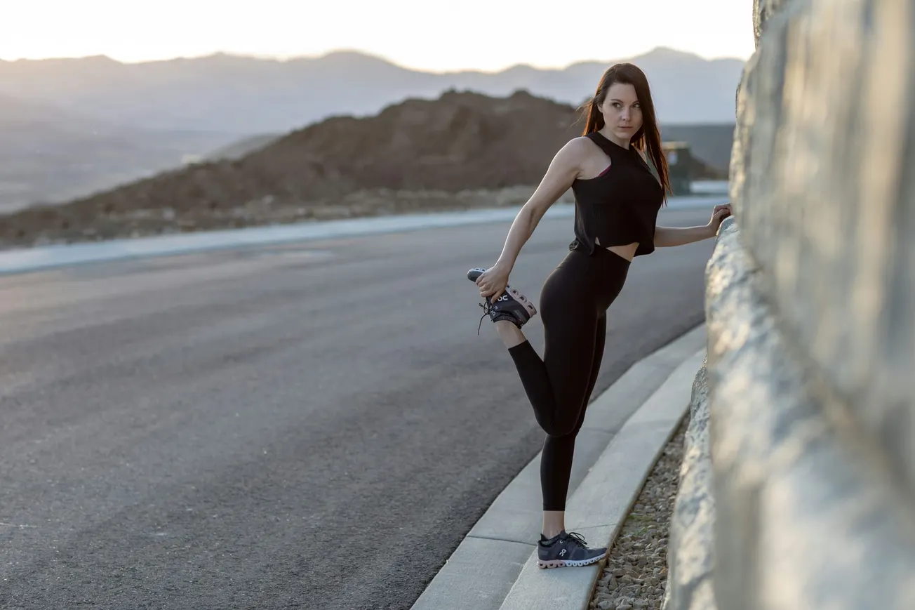 Woman stretching on a roadside near a stone wall, wearing black athletic wear. The scene is set against a backdrop of mountains at sunset.