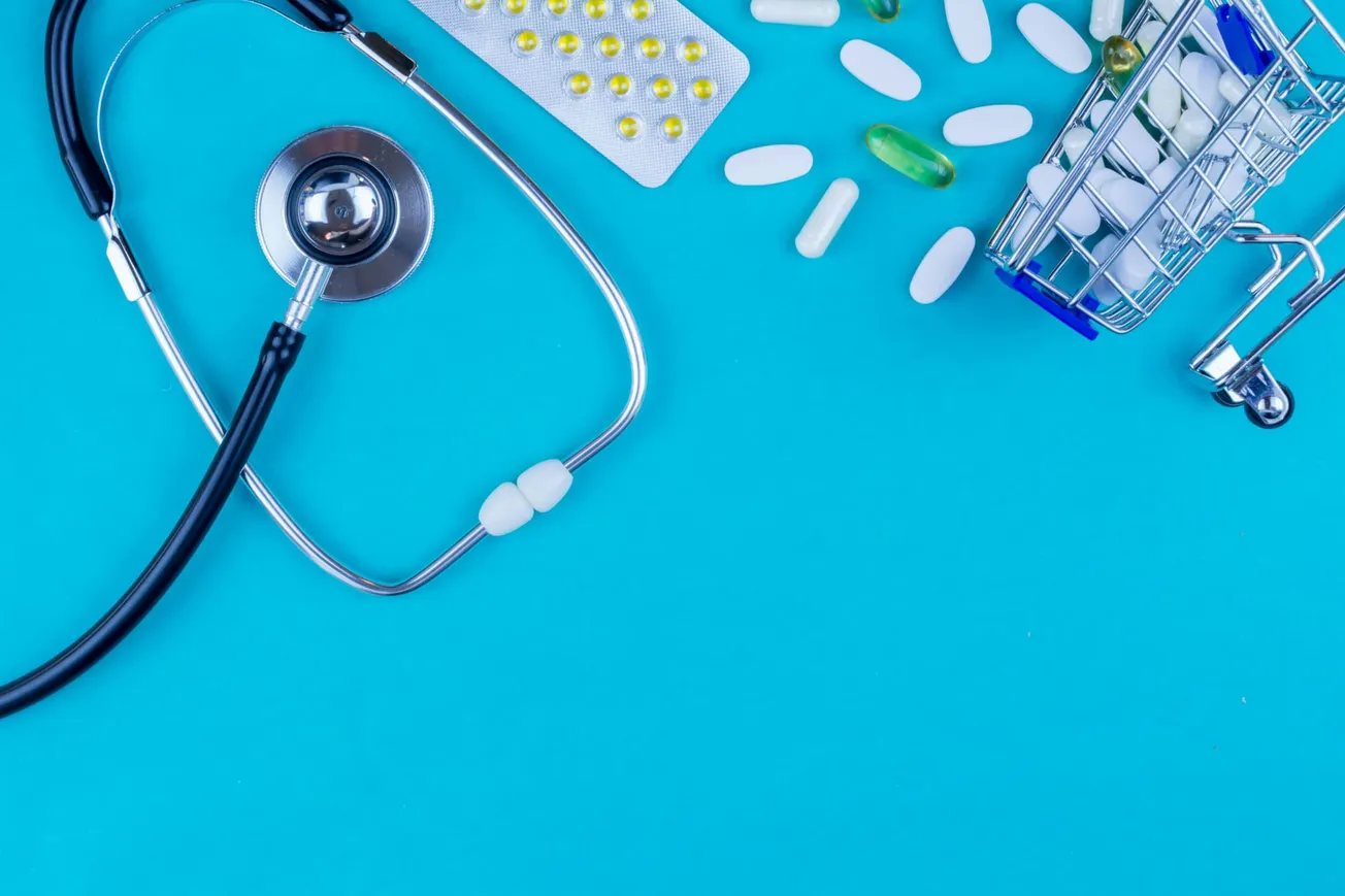 Stethoscope, pill blister pack, scattered capsules, and a miniature shopping cart on a blue background. Represents healthcare and medicine.
