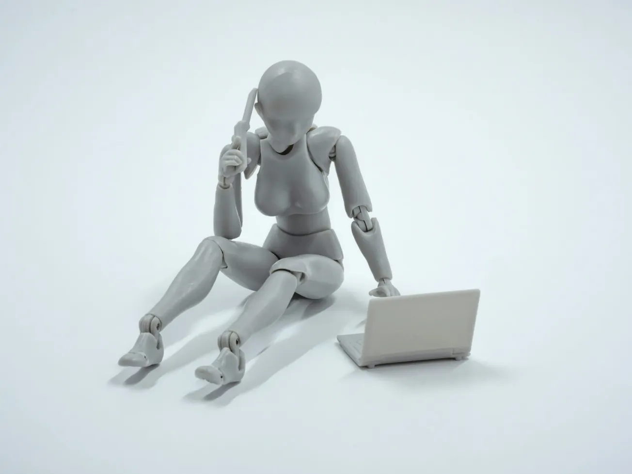 A gray humanoid figure sits on the floor, looking thoughtful with a hand to its head, while using a small laptop, set against a plain white background.