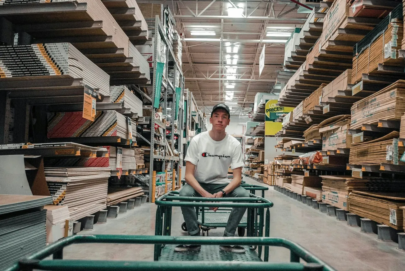 Man sitting on a flatbed cart in a hardware store aisle, surrounded by stacked wooden boards and building supplies, wearing a white T-shirt and a cap.