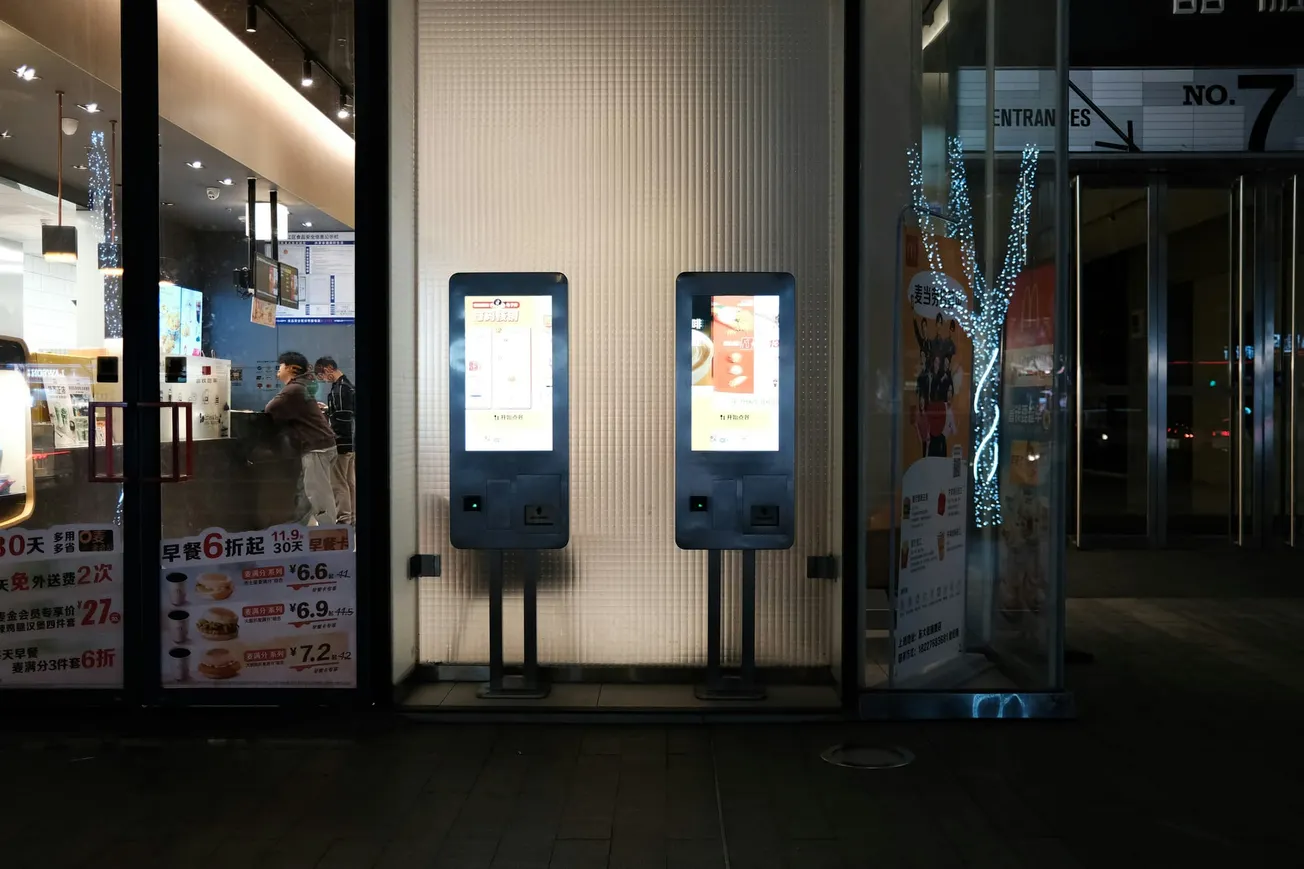 Two illuminated touchscreen kiosks stand outside a shop at night. Inside, people are visible through the glass, and a small tree wrapped in fairy lights adds brightness.