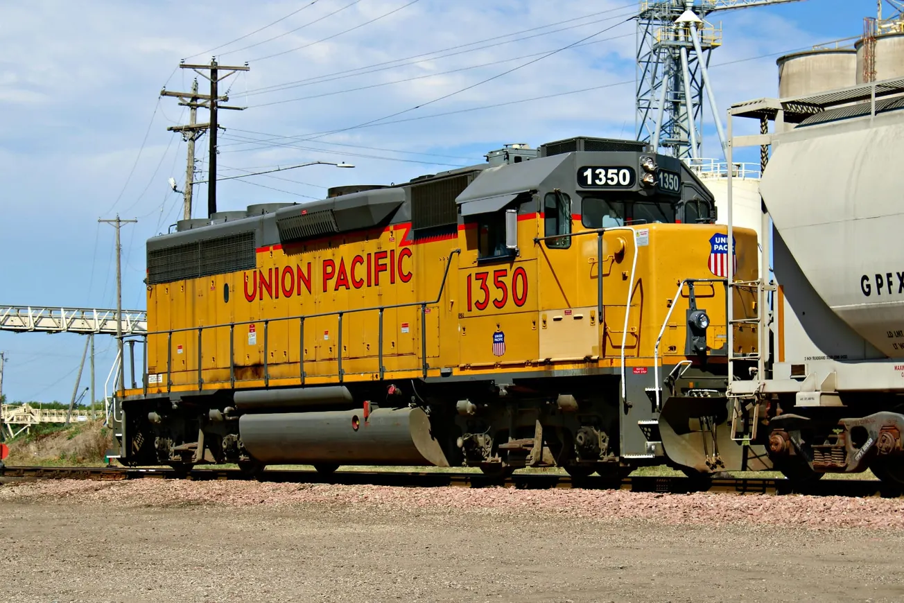 A yellow Union Pacific locomotive, numbered 1350, stands on gravel tracks beside an industrial area. Overhead power lines and a blue sky are in the background.