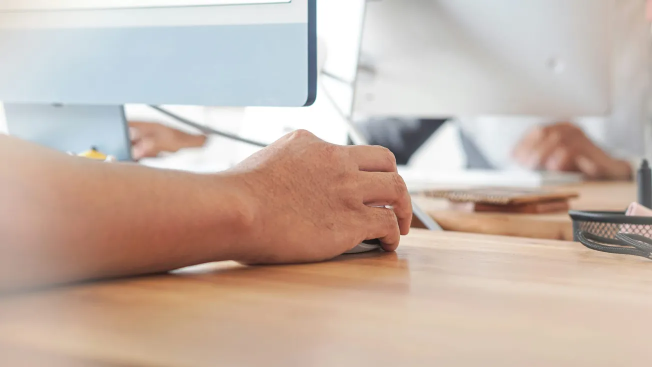 A person's hand using a computer mouse on a wooden desk, with two monitors in the background. The setting is an office, conveying focus and productivity.
