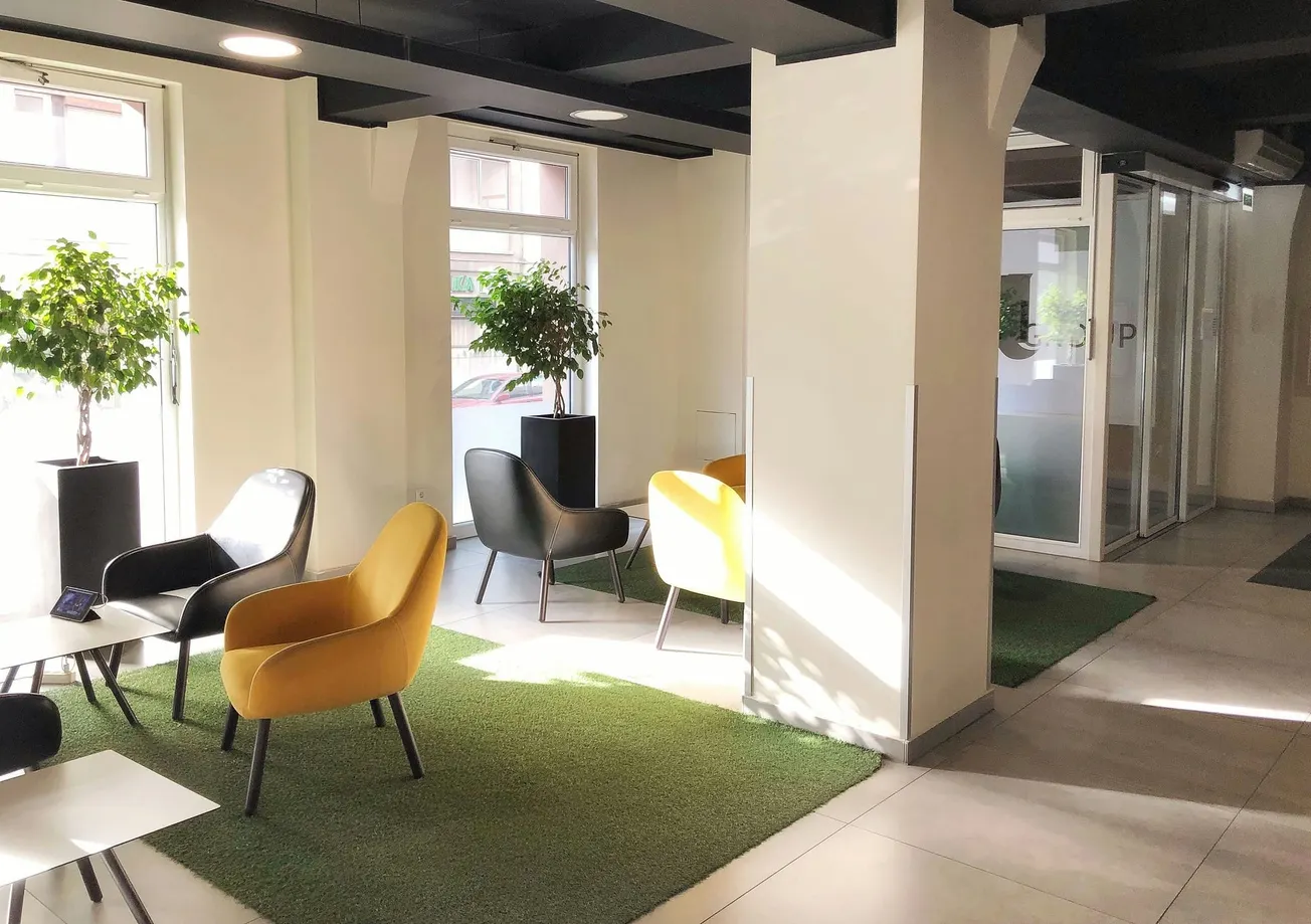 Modern lounge with yellow and black chairs on green rugs, natural light streaming through windows. Potted plants add a fresh touch, creating a calm ambiance.