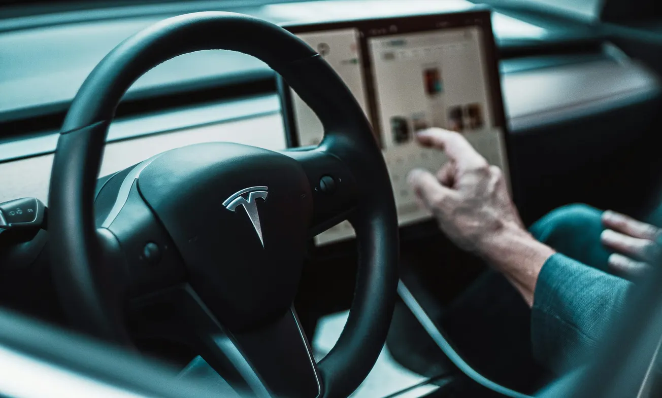 Close-up of a Tesla steering wheel and touchscreen. A person interacts with the display, suggesting modern technology and innovation inside the vehicle.