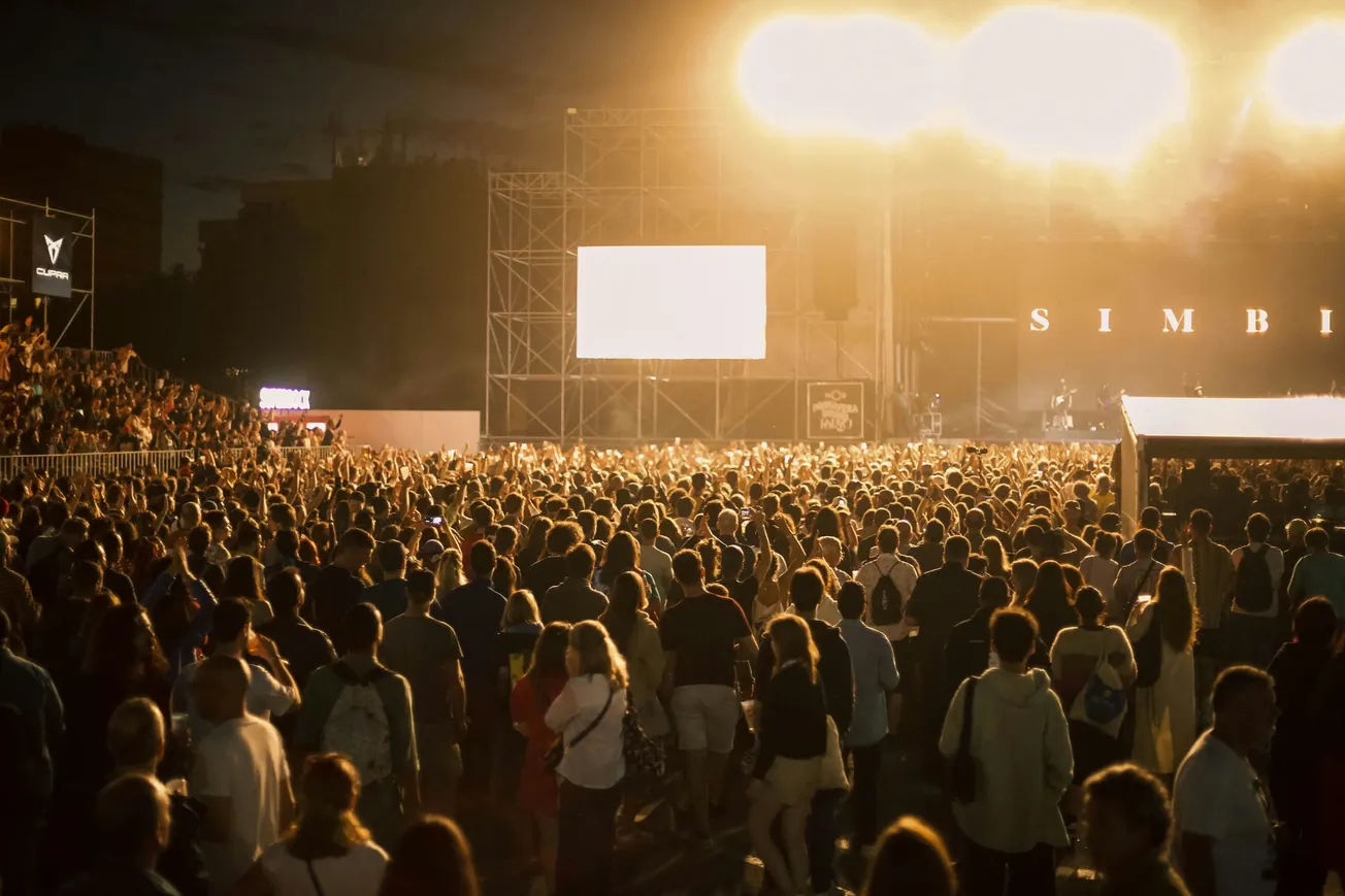 A large crowd enjoys an outdoor concert at night, with bright lights illuminating the stage. Audience members are standing closely, creating a lively atmosphere.