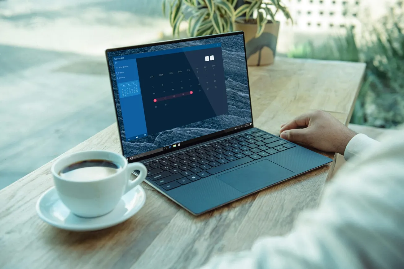 Person working on a laptop displaying a calendar on a wooden table with a cup of coffee and a plant. The ambiance feels calm and productive.