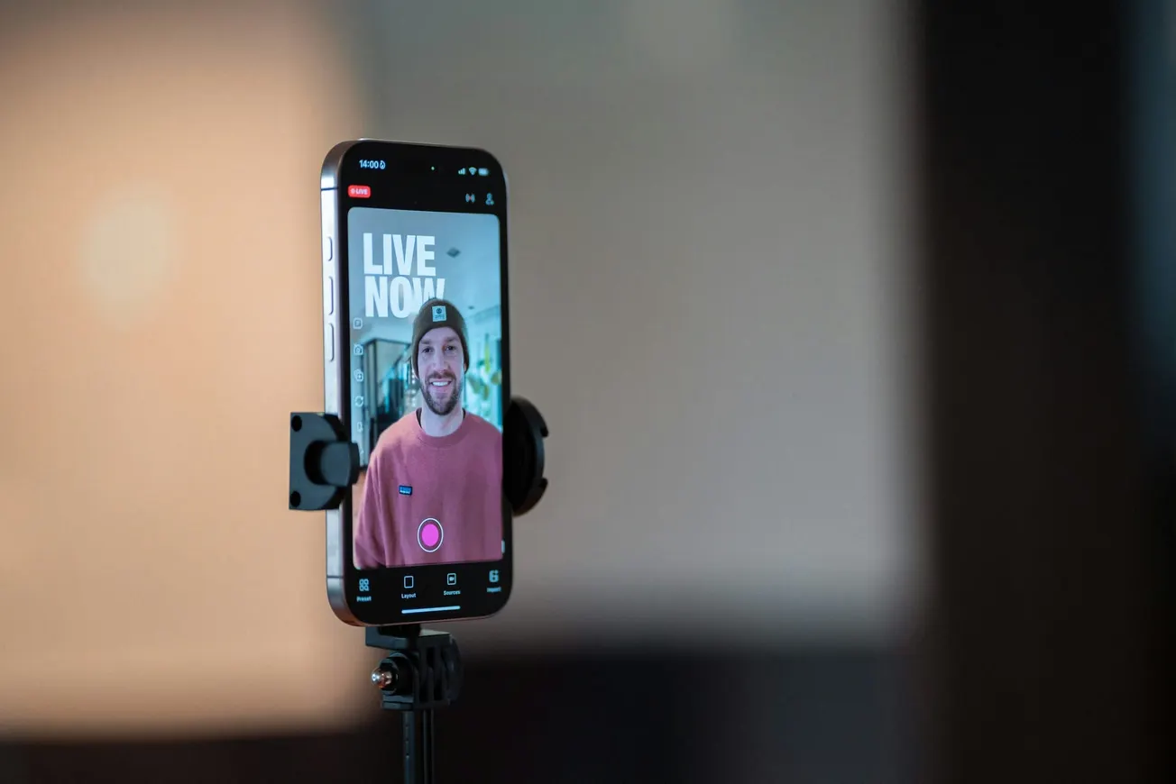 Smartphone on tripod showing a live stream of a smiling person in a beanie and pink sweater, with "LIVE NOW" on the screen, creating a casual, engaging vibe.