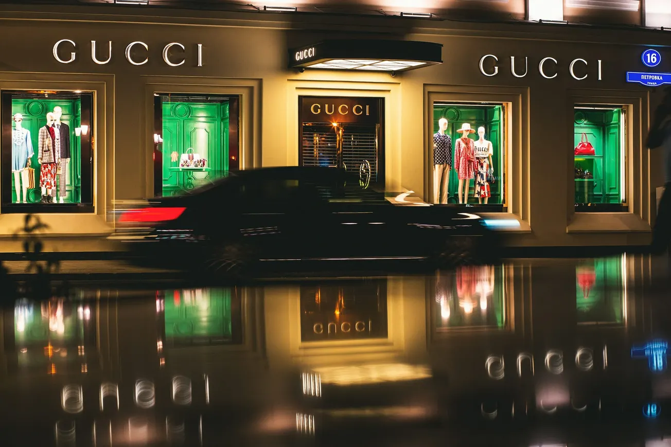 Alt text: "Night view of a Gucci store with lit windows displaying mannequins in colorful outfits. A blurred black car speeds by, reflecting on a wet street, creating a vibrant urban scene."