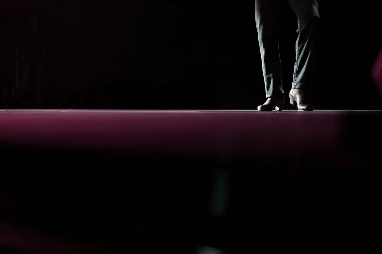 A person stands on a dimly lit stage, only their lower legs and shoes visible. The atmosphere is moody with dark surroundings and a subtle spotlight.