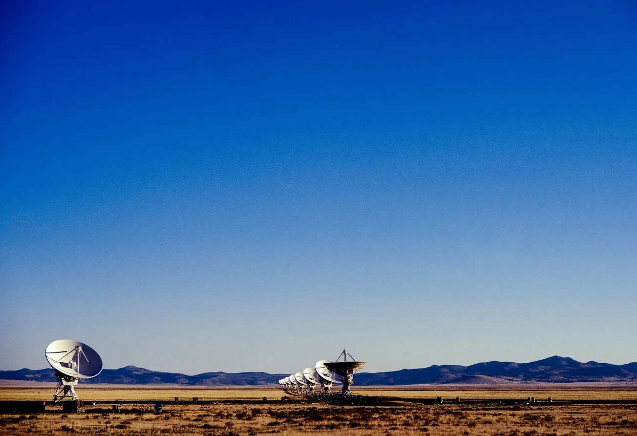 Row of large white radio telescopes in a vast desert landscape under a clear blue sky, conveying a sense of exploration and isolation.