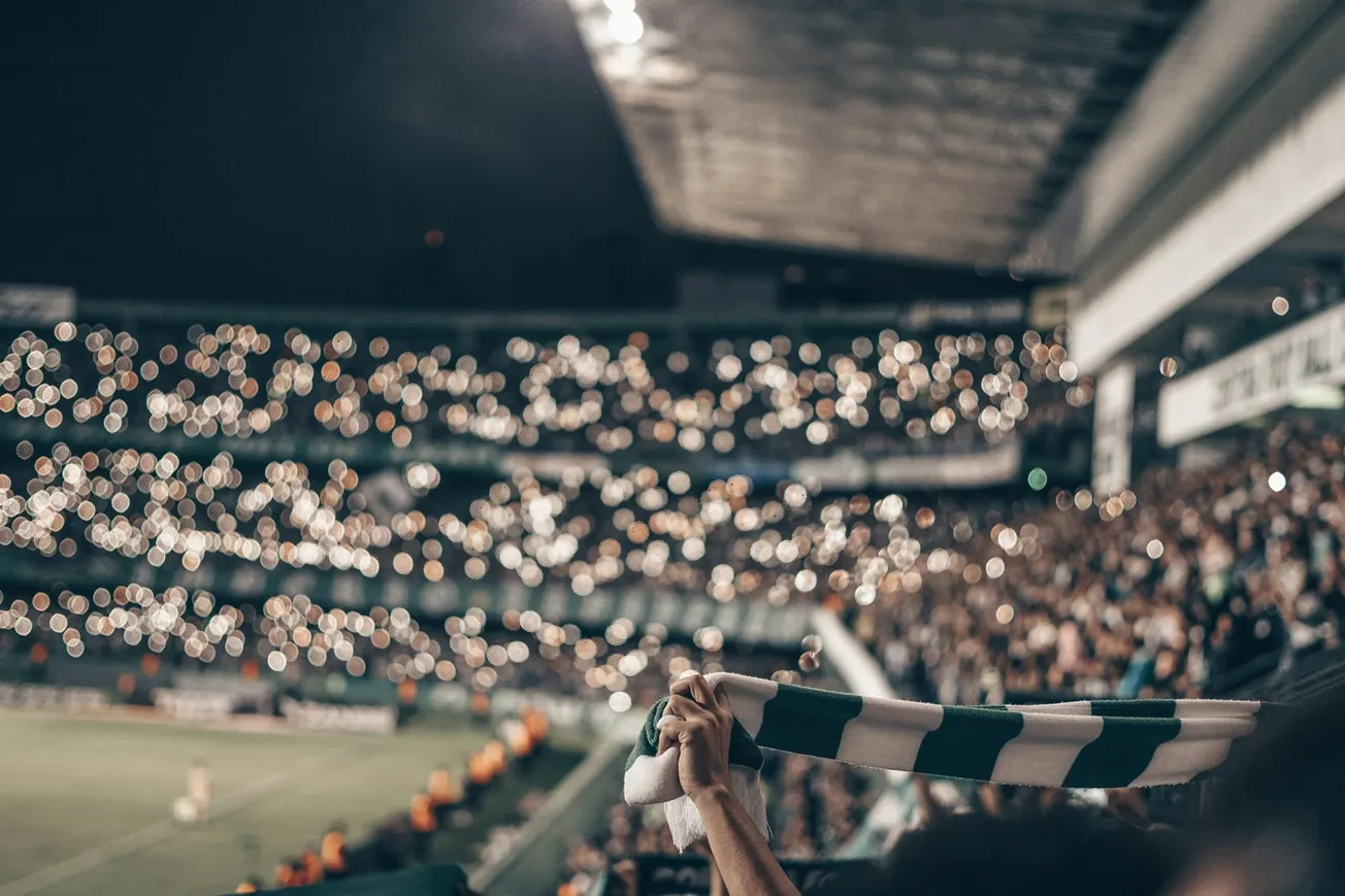 Fans in a dimly lit stadium hold up a green and white scarf, while numerous lights sparkle across the stands, creating a festive, unified atmosphere.