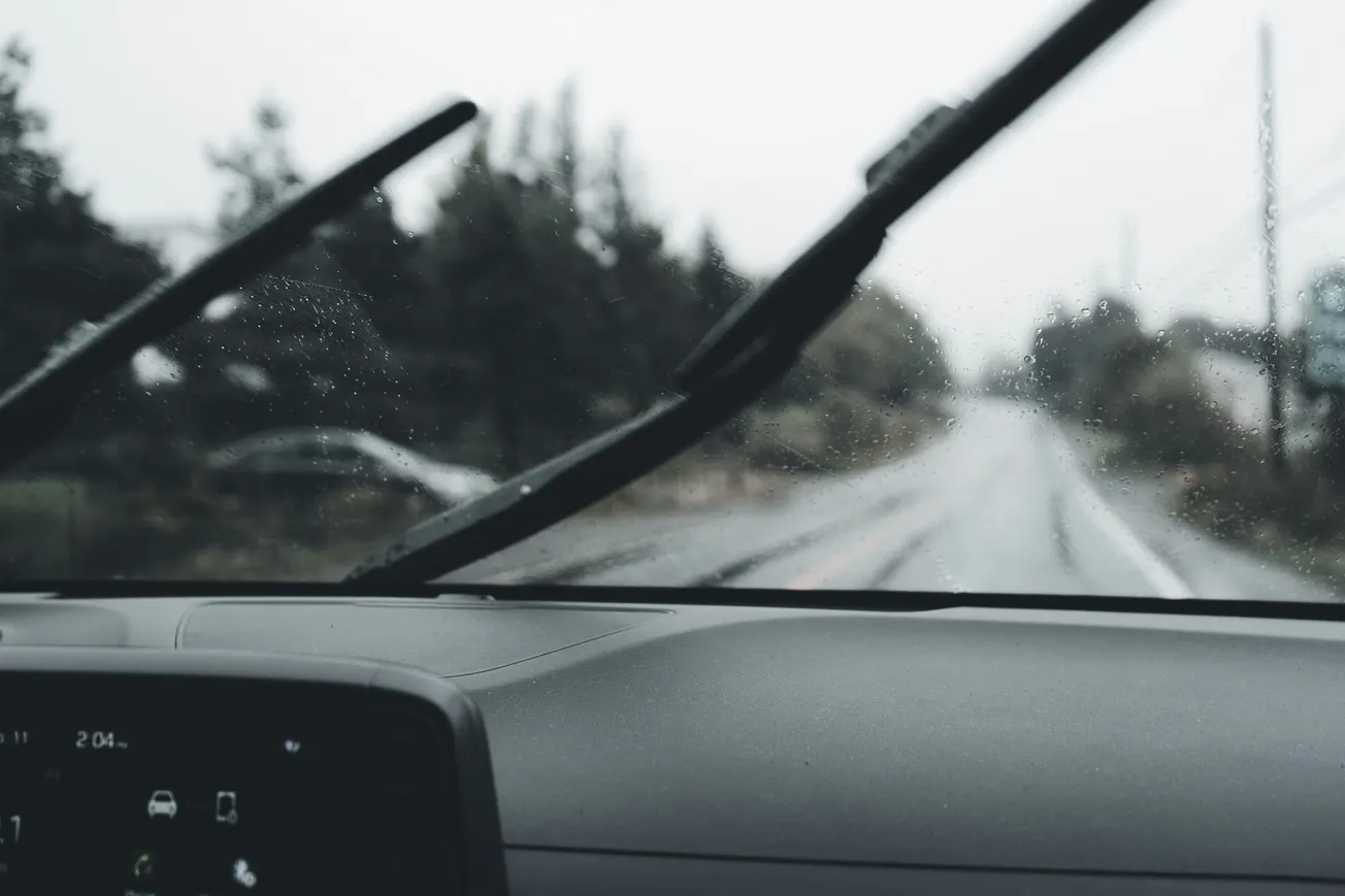View from inside a car on a rainy day, showing windshield wipers in motion and a blurred, wet road ahead, creating a calm, subdued mood.