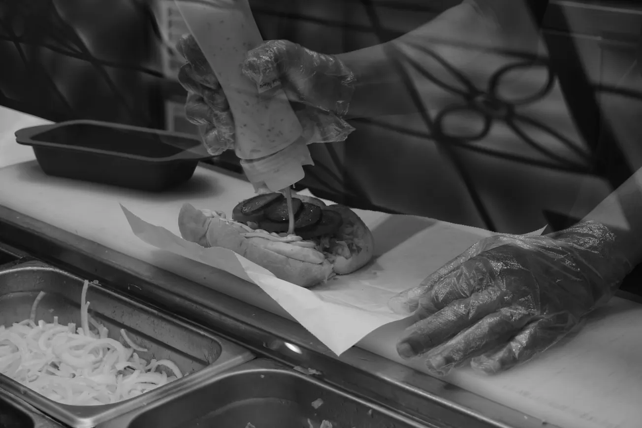 Person in gloves applies sauce on a sandwich with pickles in a kitchen setting. Ingredients like onions are visible in metal containers below.