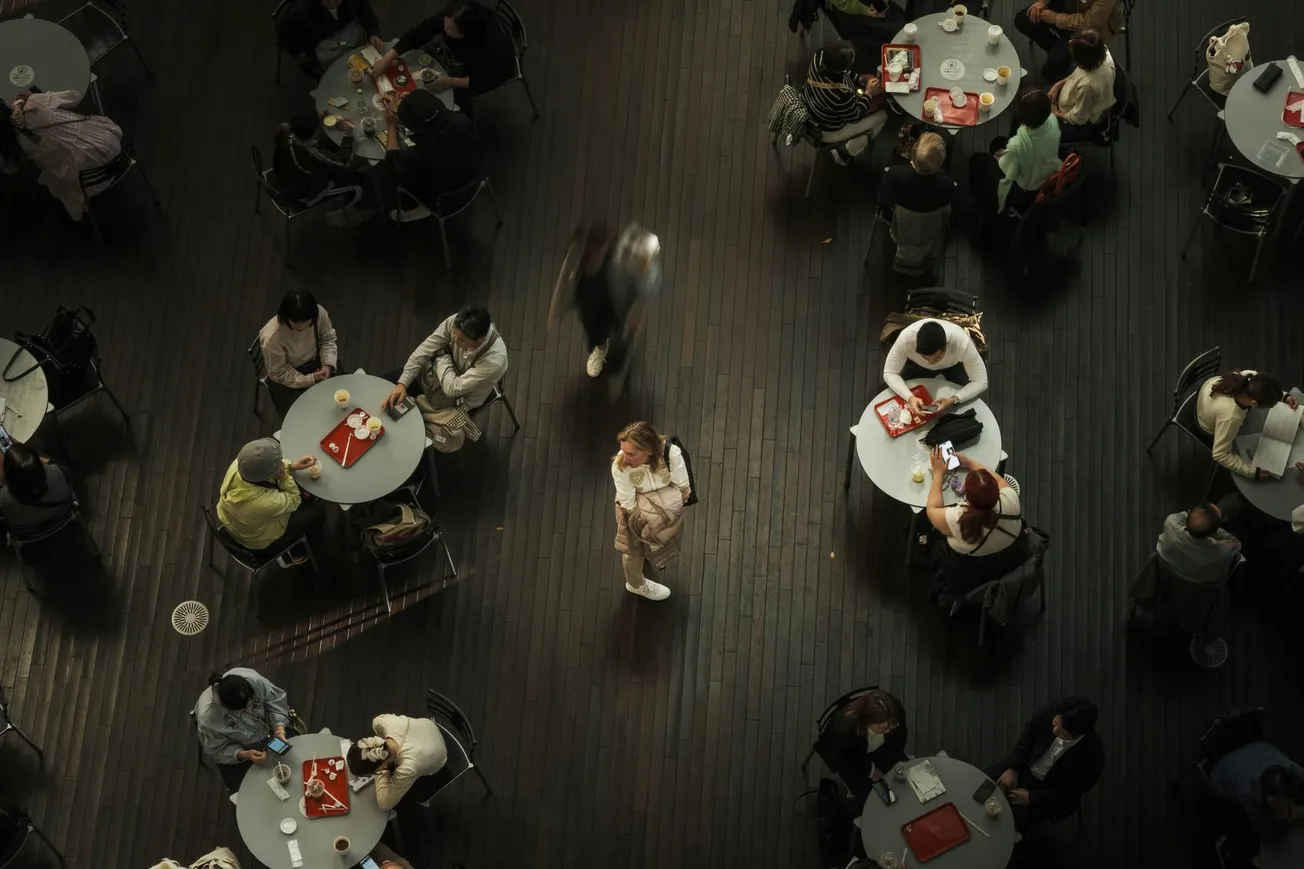 Aerial view of a busy cafe with circular tables on a dark wood floor. Patrons eat and converse, while a person stands alone in the center, creating contrast.