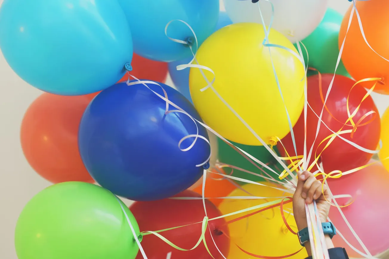 A vibrant cluster of colorful balloons, including blue, green, yellow, and red, are held by a hand. The scene conveys a joyful celebration.