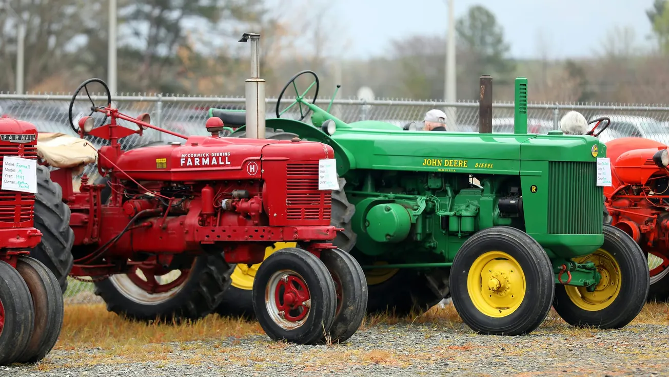 Three vintage tractors are parked on gravel: a red Farmall, a green John Deere, and an orange tractor. Overcast sky and trees in the background.