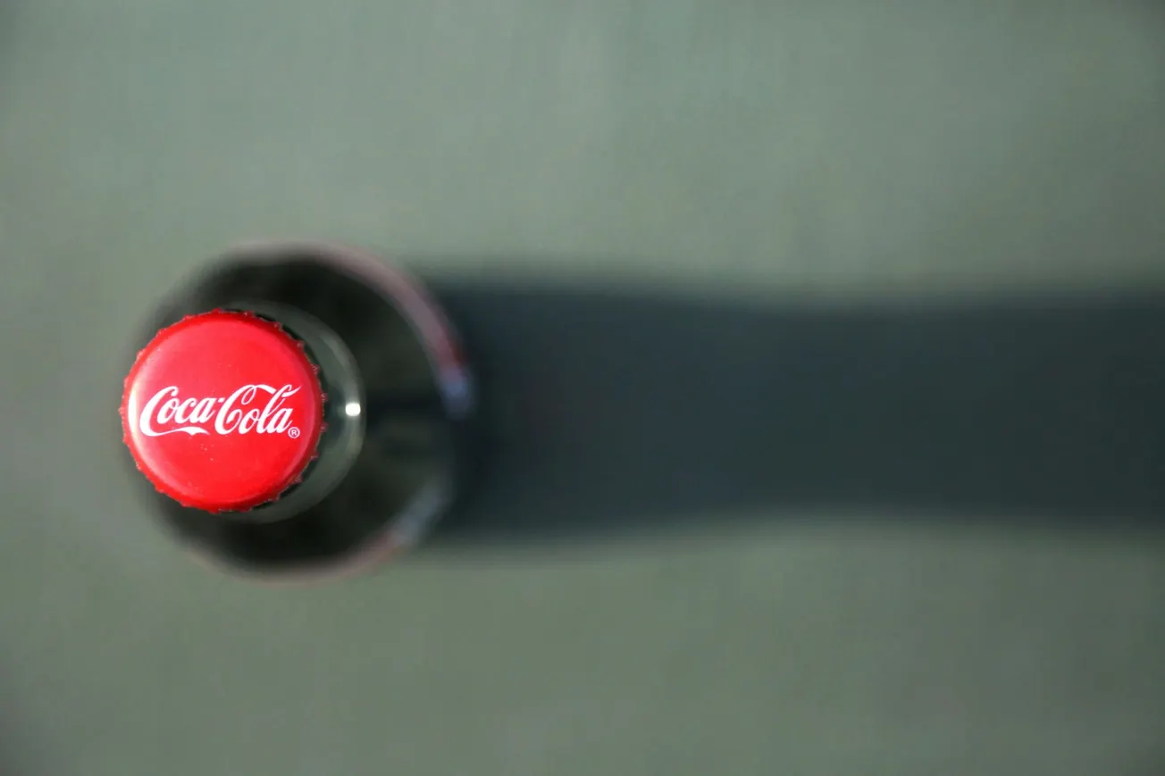 Overhead view of a Coke bottle with a red cap showing the Coca-Cola logo. The bottle casts a long shadow on a gray surface, conveying a clean, minimalist feel.