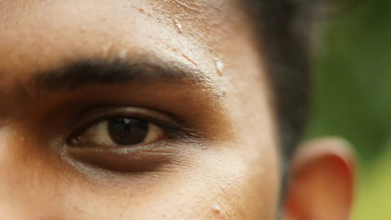 Close-up of a person's eye and part of their face, showing beads of sweat on their forehead. The background is blurred green, conveying a sense of warmth.