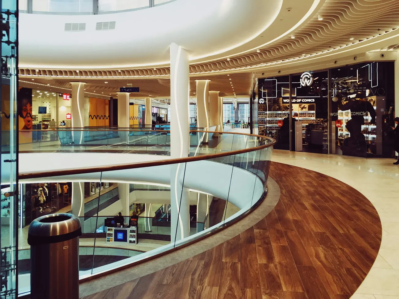 Modern indoor shopping mall with elegant curved wooden flooring and glass railings. Stores line the walls under soft, wavy ceiling lights.