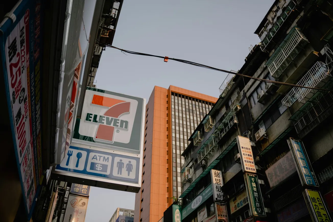 Street scene with a 7-Eleven sign amidst tall buildings, some with signs in various languages. The sky is clear, creating an urban ambiance.