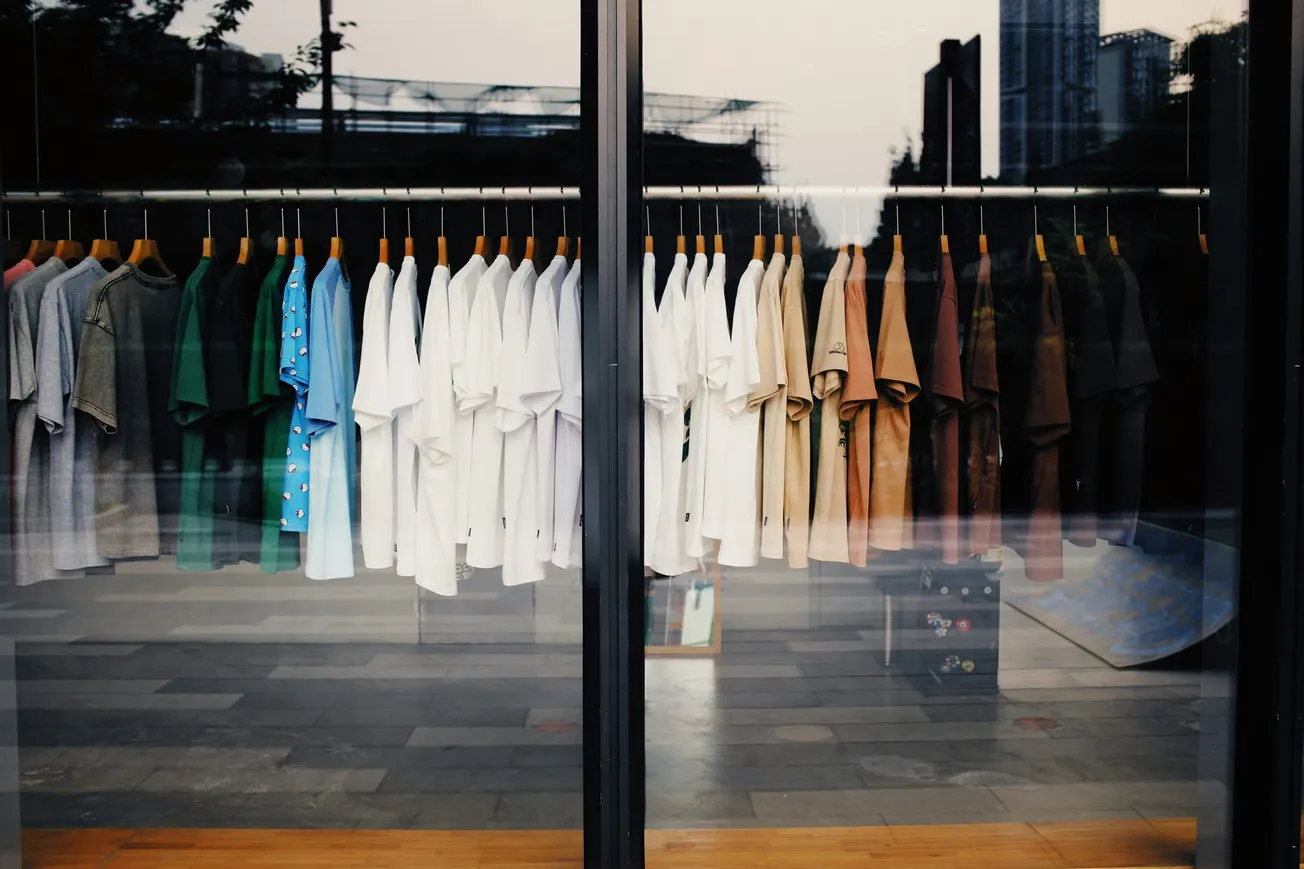 A row of colorful T-shirts, including green, blue, white, beige, and brown, hang neatly in a store window. The urban background is reflected on the glass.
