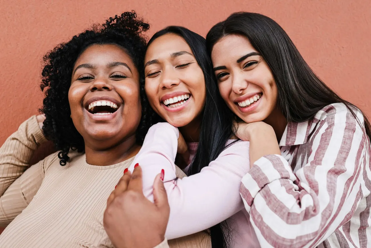 Three friends stand closely, smiling joyfully against a warm, orange wall. Their expressions convey happiness and a sense of camaraderie.