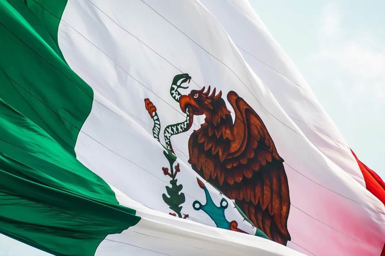Close-up of the Mexican flag waving, showing the green, white, and red stripes with a prominent eagle holding a snake on a cactus in the center.