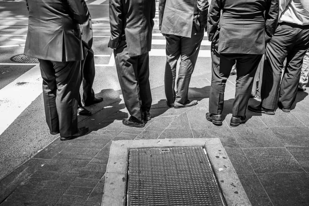 A group of men in suits stand at a street corner, captured from the waist down. The black and white image emphasizes business formality and urban life.