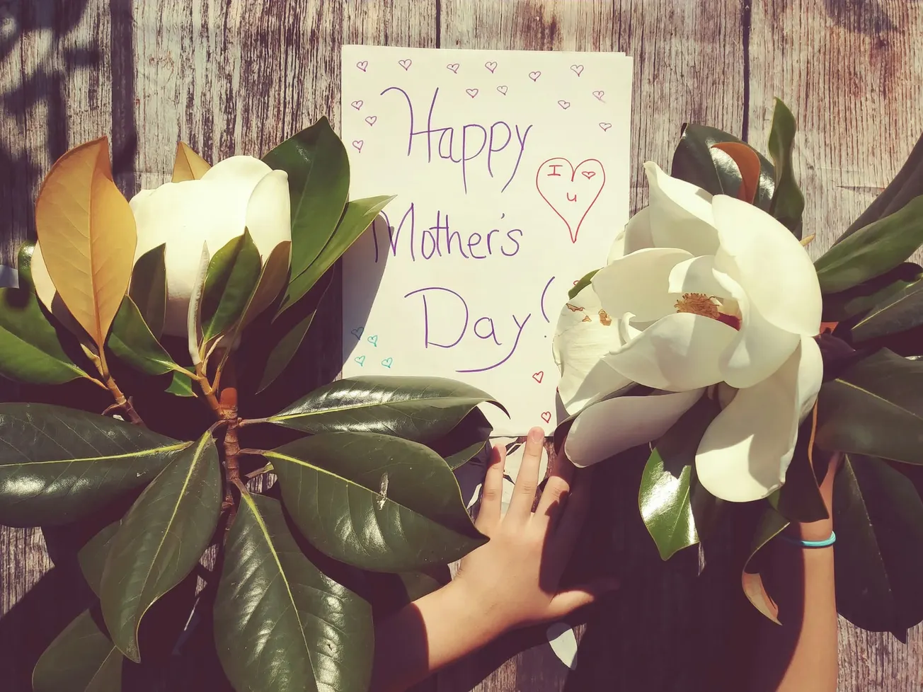 A child places two magnolia flowers on a wooden table, flanking a handmade card that reads "Happy Mother's Day!" with colorful hearts.
