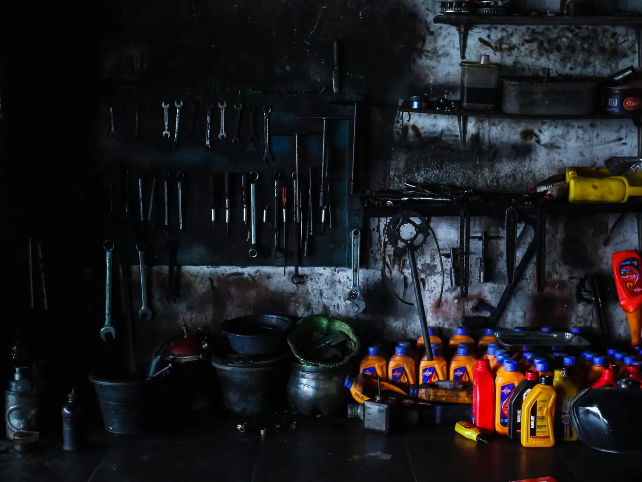 Dimly lit garage wall with hanging tools and a cluttered shelf. Brightly colored oil containers and buckets below exude a rustic, industrious vibe.