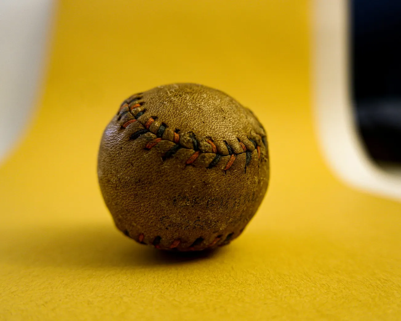 A worn brown baseball with visible red and black stitching sits on a yellow surface. The background is a soft gradient from yellow to white.