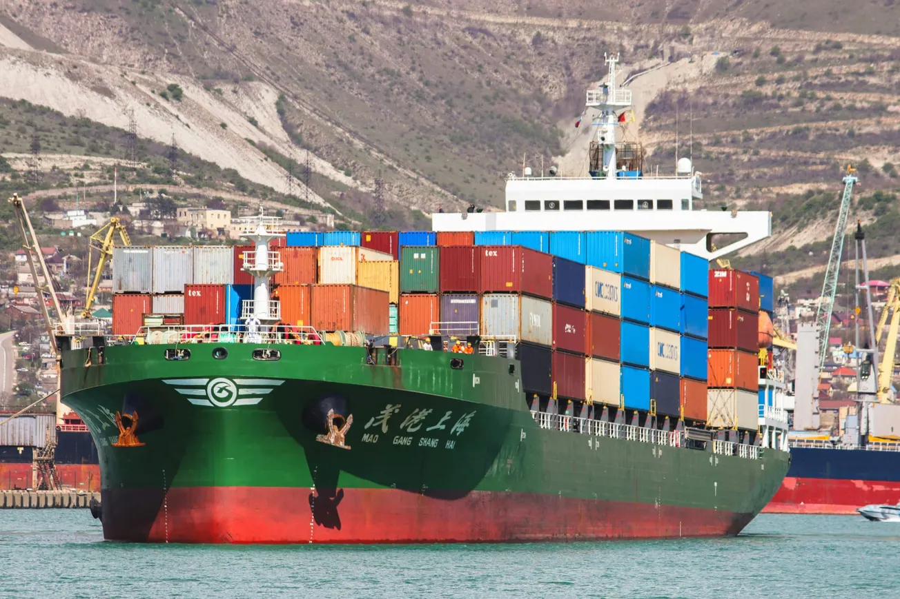 A large green cargo ship is loaded with colorful shipping containers in a busy port. Background features hilly terrain and industrial structures.
