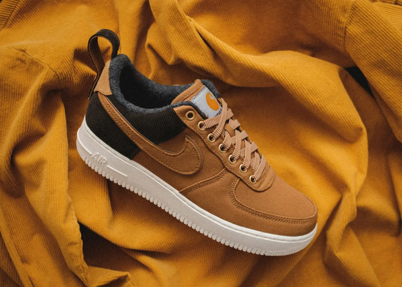 Sneaker with brown fabric upper and white sole, featuring a swoosh logo, lace-up design, placed on matching brown textured fabric, creating a warm tone.