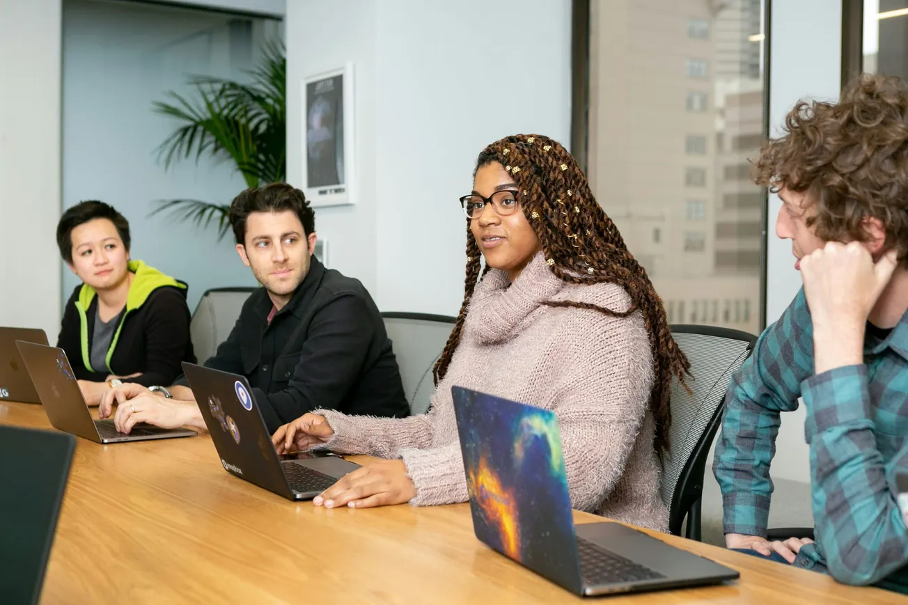 Four people sit at a conference table with laptops, engaged in discussion. A bright, modern office setting with a large window and a plant.