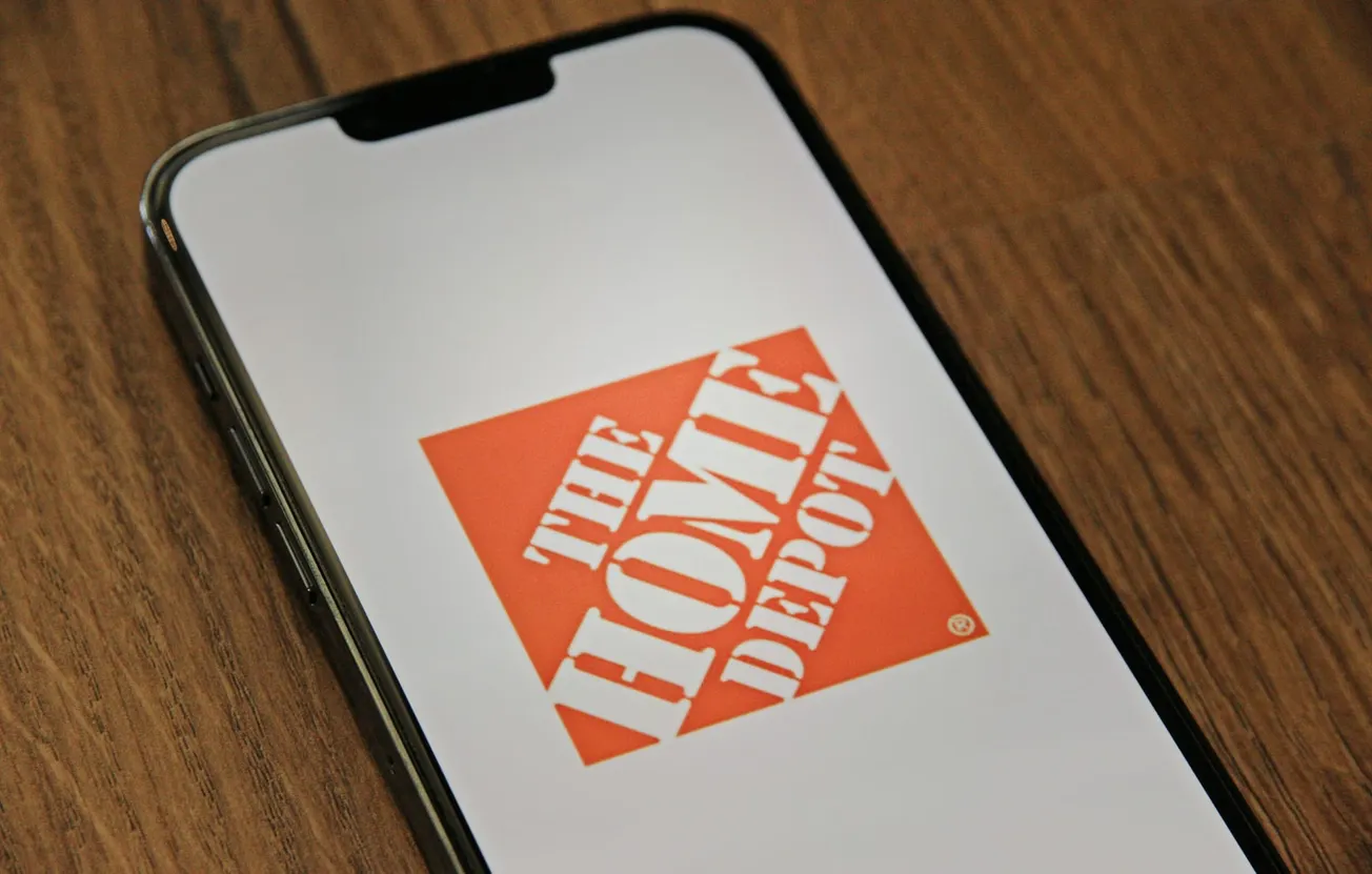 Smartphone on a wooden surface displaying The Home Depot logo. The orange and white logo is bold and prominent, suggesting a focus on home improvement.