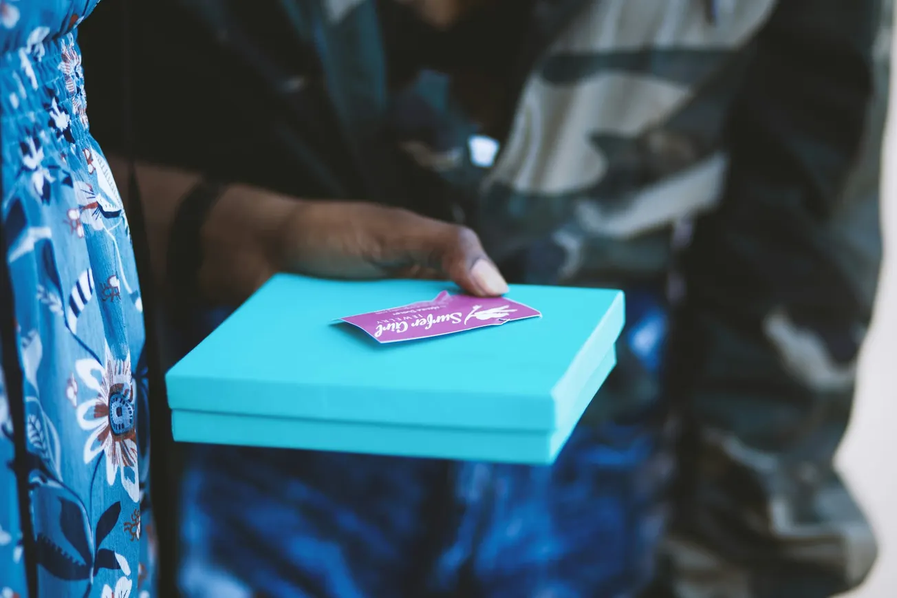 A person holds a turquoise gift box with a purple "Surprise Gift" card. They wear a floral dress, and another person wears a camo jacket, suggesting a thoughtful exchange.