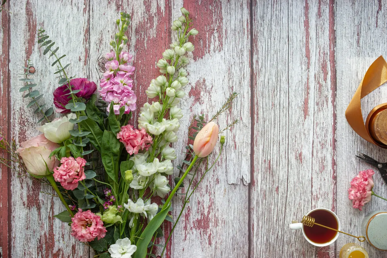 A bouquet of pink, white, and purple flowers lies on a rustic, weathered wooden surface. A teacup with honey and a ribbon are on the right. Serene and cozy.