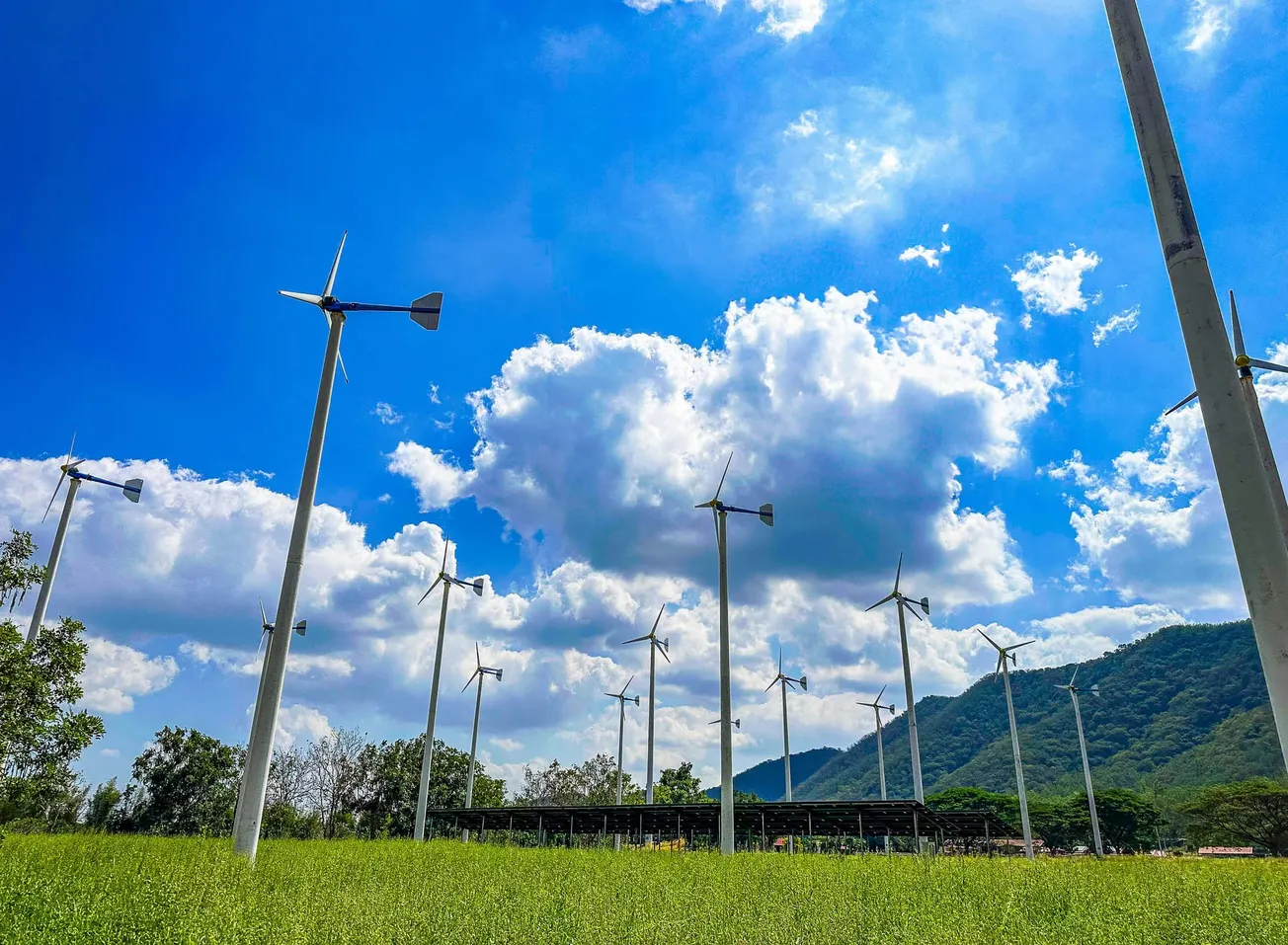 Wind turbines on a lush green field against a bright blue sky with fluffy clouds. A mountainous backdrop adds depth to the serene, sustainable energy scene.