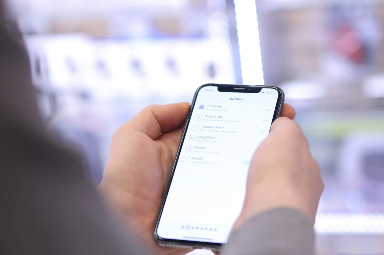 A person holds a smartphone displaying a menu for button settings, including "Proximity" and "Face ID." The background is softly blurred, evoking a tech-focused ambiance.