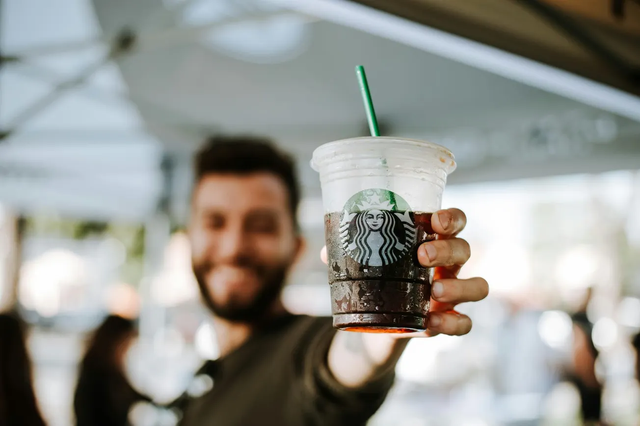 A smiling man holds out a cold Starbucks drink with ice, the brand's logo visible. The background is a blurred outdoor café setting, conveying a cheerful vibe.