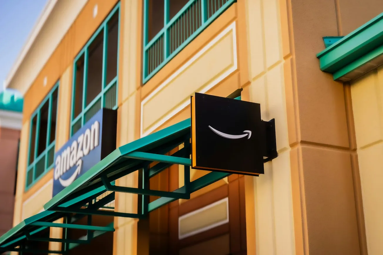 Alt text: "Exterior of an Amazon store with a tan and green facade. A black sign featuring the Amazon smile logo hangs above a teal awning. Bright, inviting atmosphere."