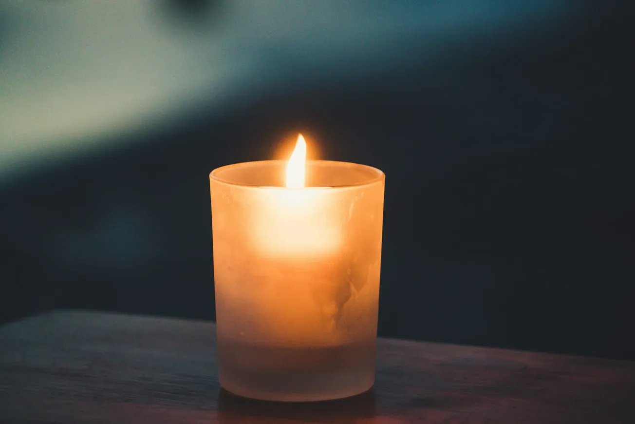 A single lit candle in a frosted glass holder emits a warm, soft glow, set against a dark, blurred background, creating a peaceful, tranquil mood.