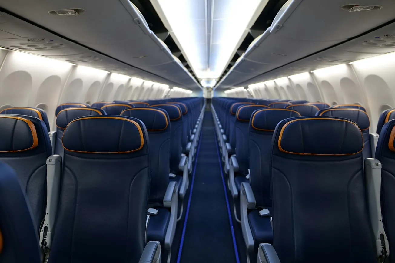 Empty airplane cabin with rows of dark blue leather seats and orange trim, illuminated by overhead lighting. Aisle is centered, evoking a calm, orderly tone.
