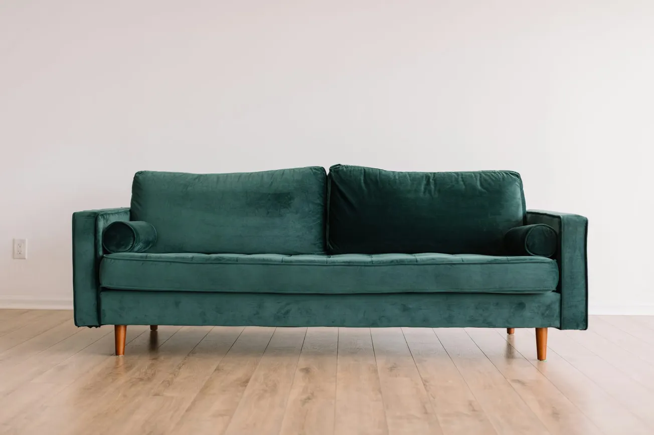 A teal velvet sofa with wooden legs is centered against a plain white wall. The room has light wood flooring, creating a minimalist, modern feel.