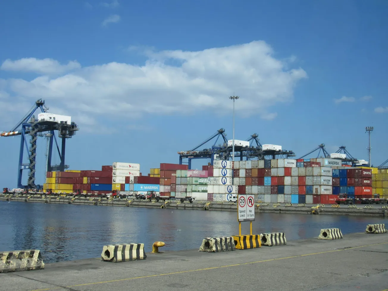 A busy port with stacked colorful shipping containers, large cranes in the background under a mostly clear blue sky, and calm water in the foreground.