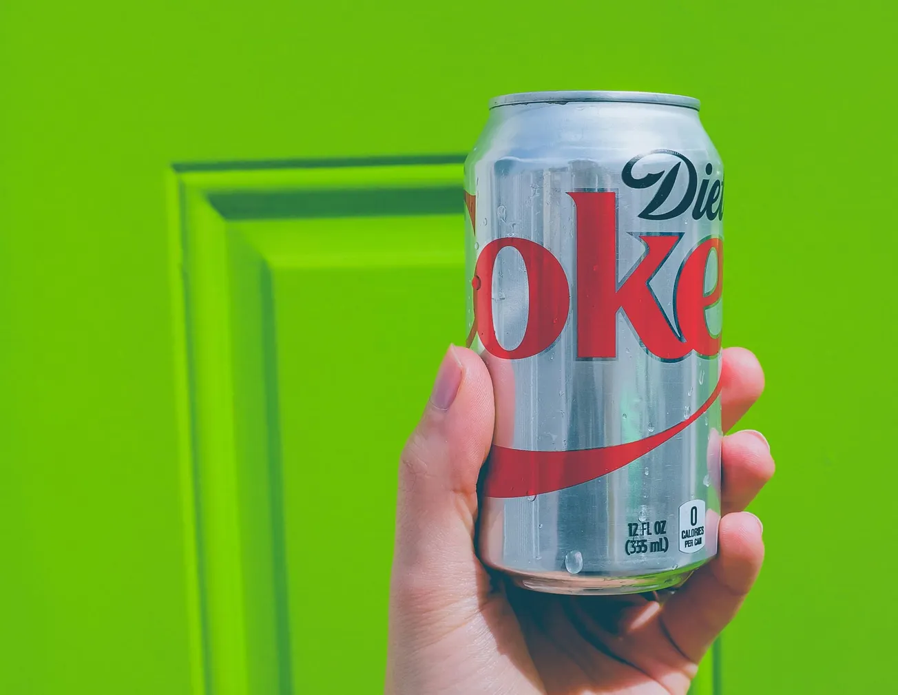 Hand holding a can of Diet Coke against a vibrant lime green background. The scene has a refreshing and lively feel.